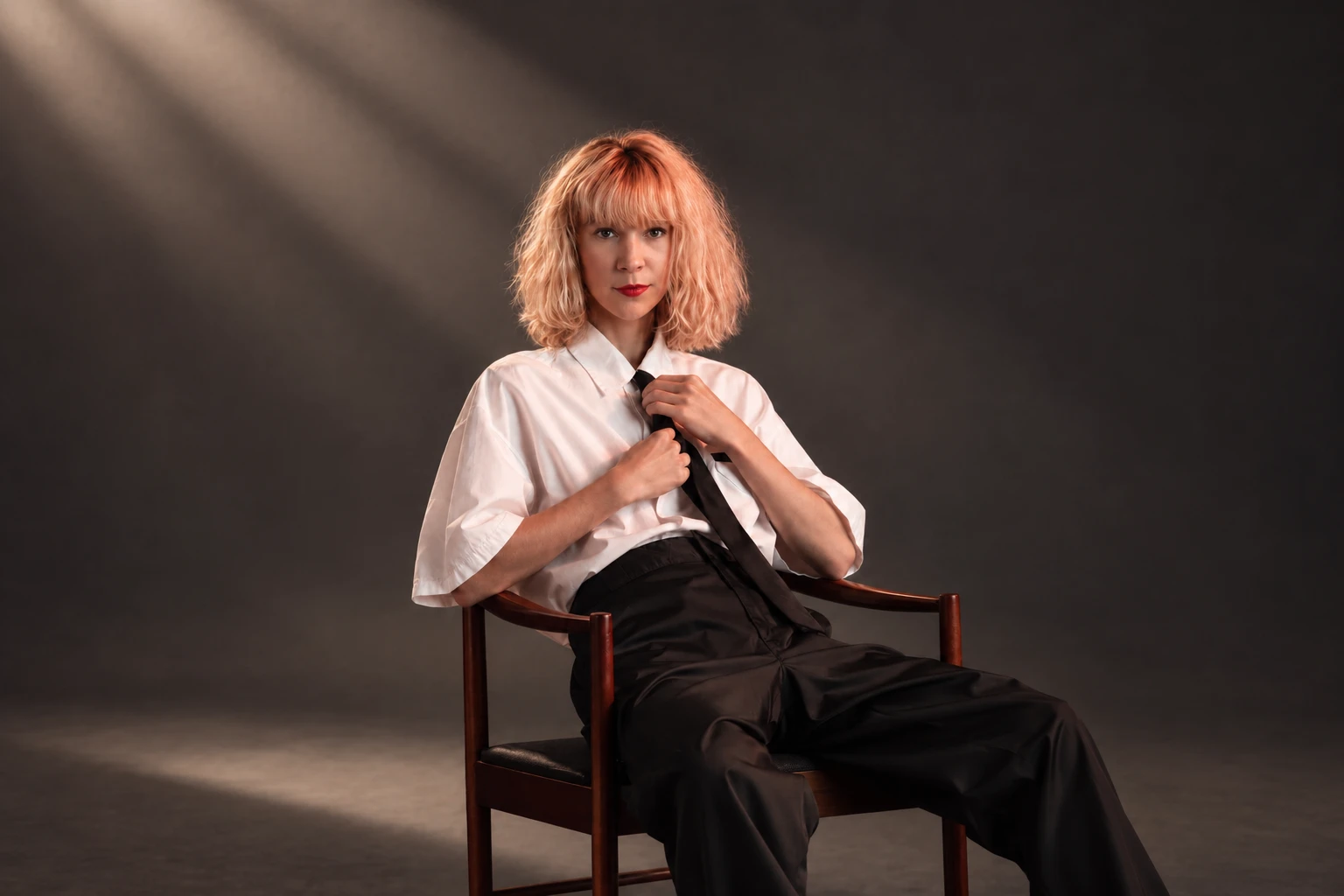 Emeraldia assise sur une chaise en bois, chemise blanche et cravate noire, dans un rai de lumière